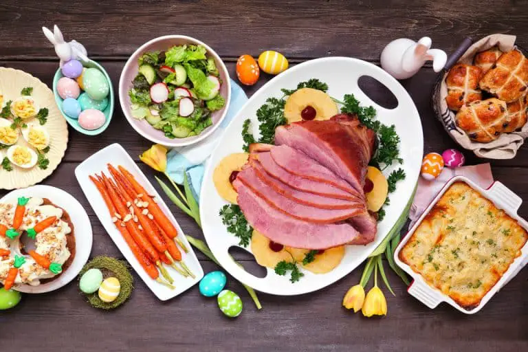 Traditional Easter ham dinner. Top down view table scene on a dark wood background. Ham, scalloped potatoes, vegetables, eggs, hot cross buns and carrot cake.