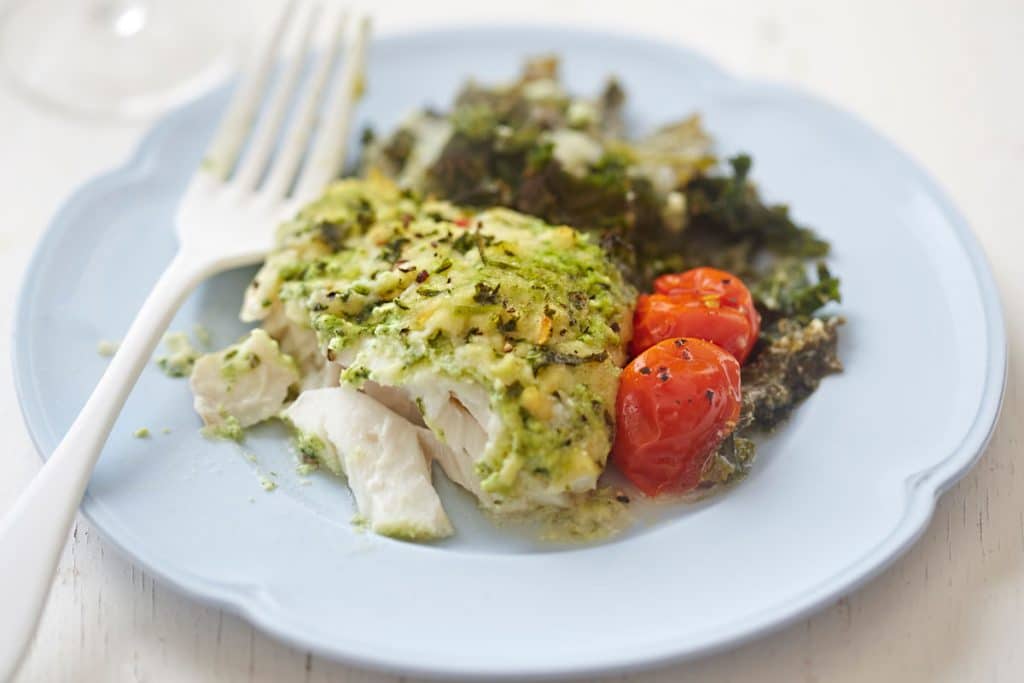 pesto baked cod with cherry tomatoes and greens on white plate