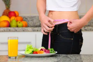 person holding pink measuring tape around their waist with glass of oj and salad on counter